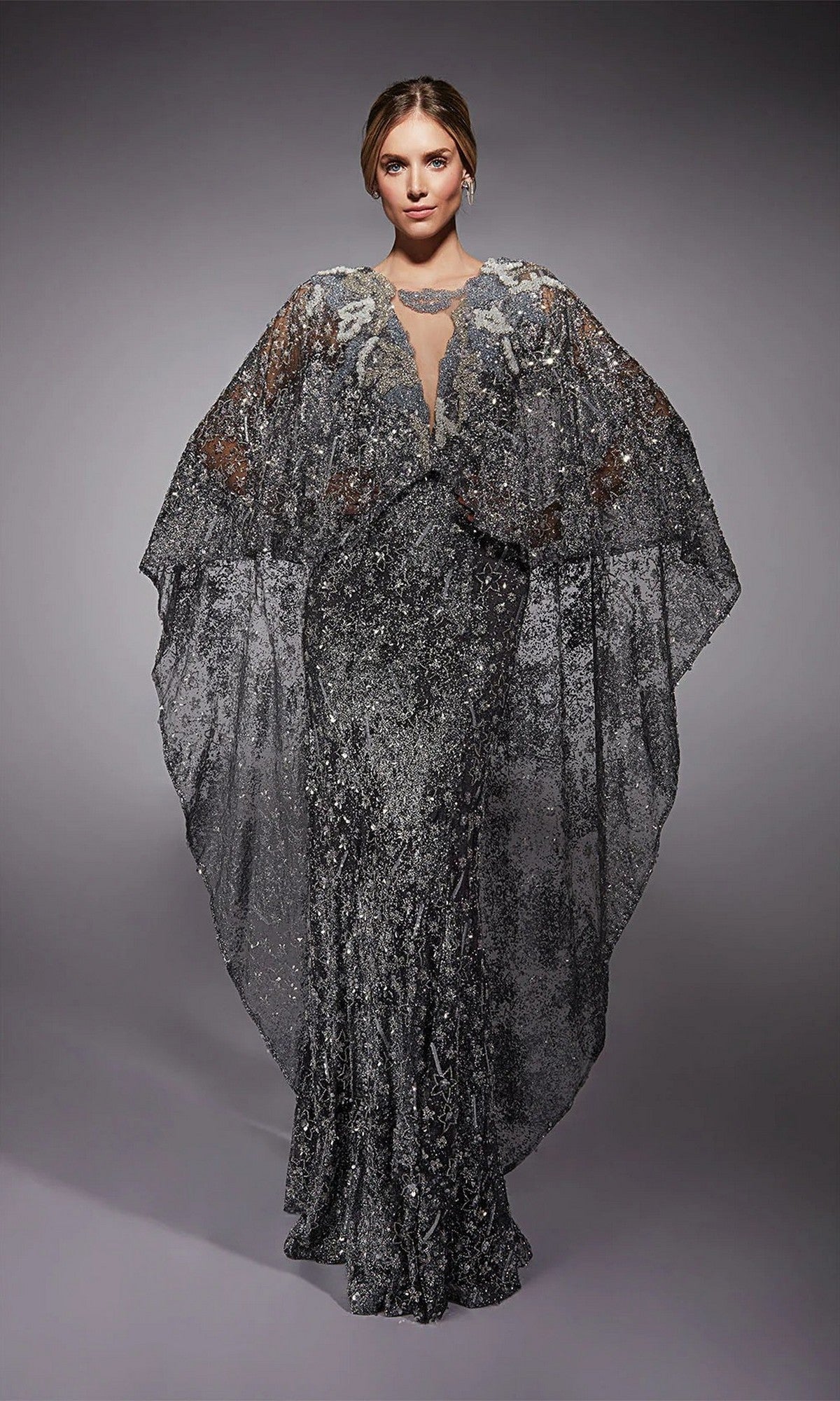 A person poses against a plain gray background wearing the Alyce 27752 long formal dress in charcoal, featuring floor-length glitter tulle and a sheer, wing-like cape.